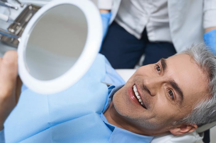 Male patient smiling in dental chair holding mirror during checkup examination.