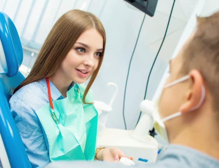 Female dentist in blue scrubs explains treatment to male patient in dental office.