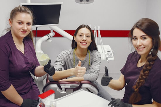 Three dental professionals in purple scrubs giving thumbs up in modern dental office treatment room.