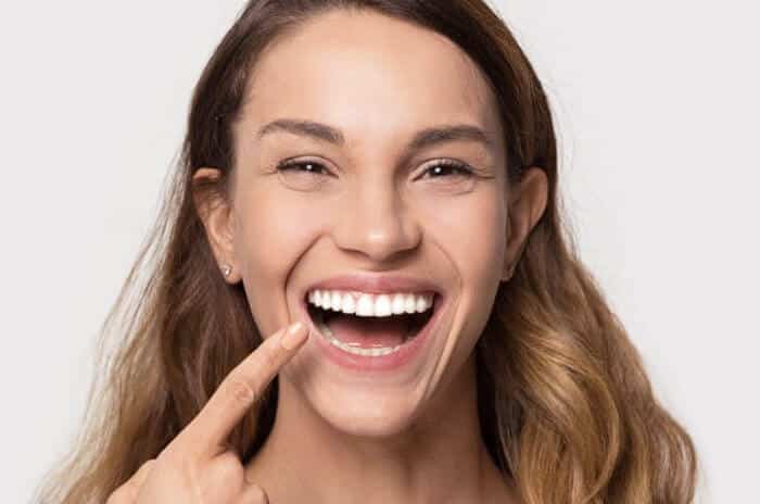 Woman smiling brightly while pointing to her teeth against neutral background.