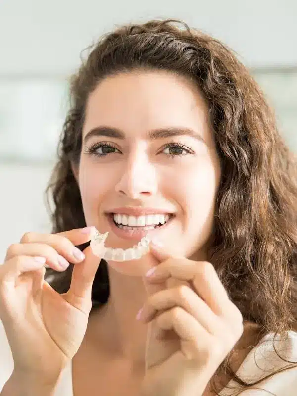 Woman holding clear dental aligners, smiling in bright clinical setting.