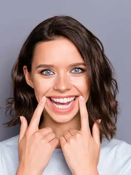Smiling woman pointing to her teeth in dental office setting against gray background.