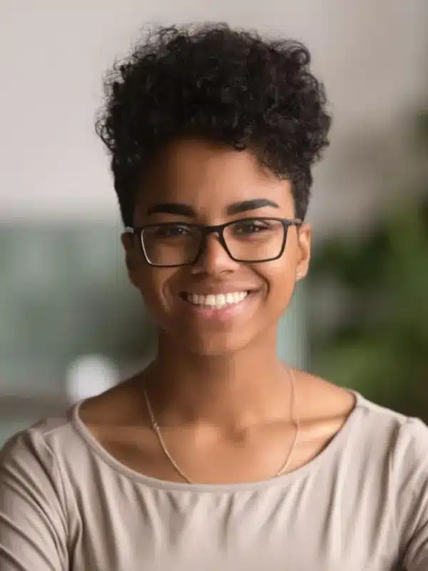 Young person wearing glasses and beige shirt smiling at camera in dental office setting.