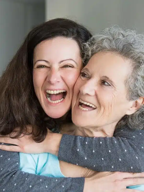 Two women smiling warmly together in an indoor setting, embracing in a close, joyful moment.