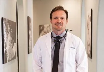 Smiling male dentist in white coat and tie standing in modern dental office hallway.