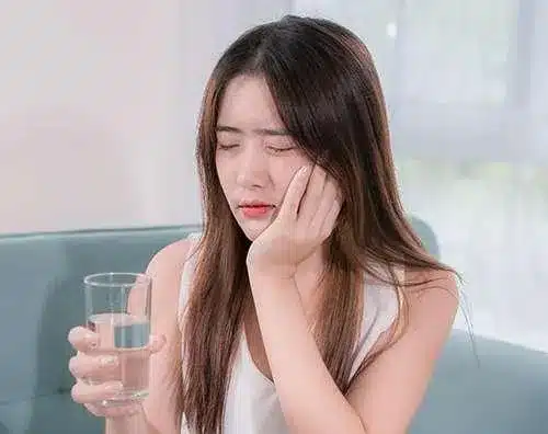 Woman holding water glass with pained facial expression, suggesting tooth sensitivity or dental discomfort.