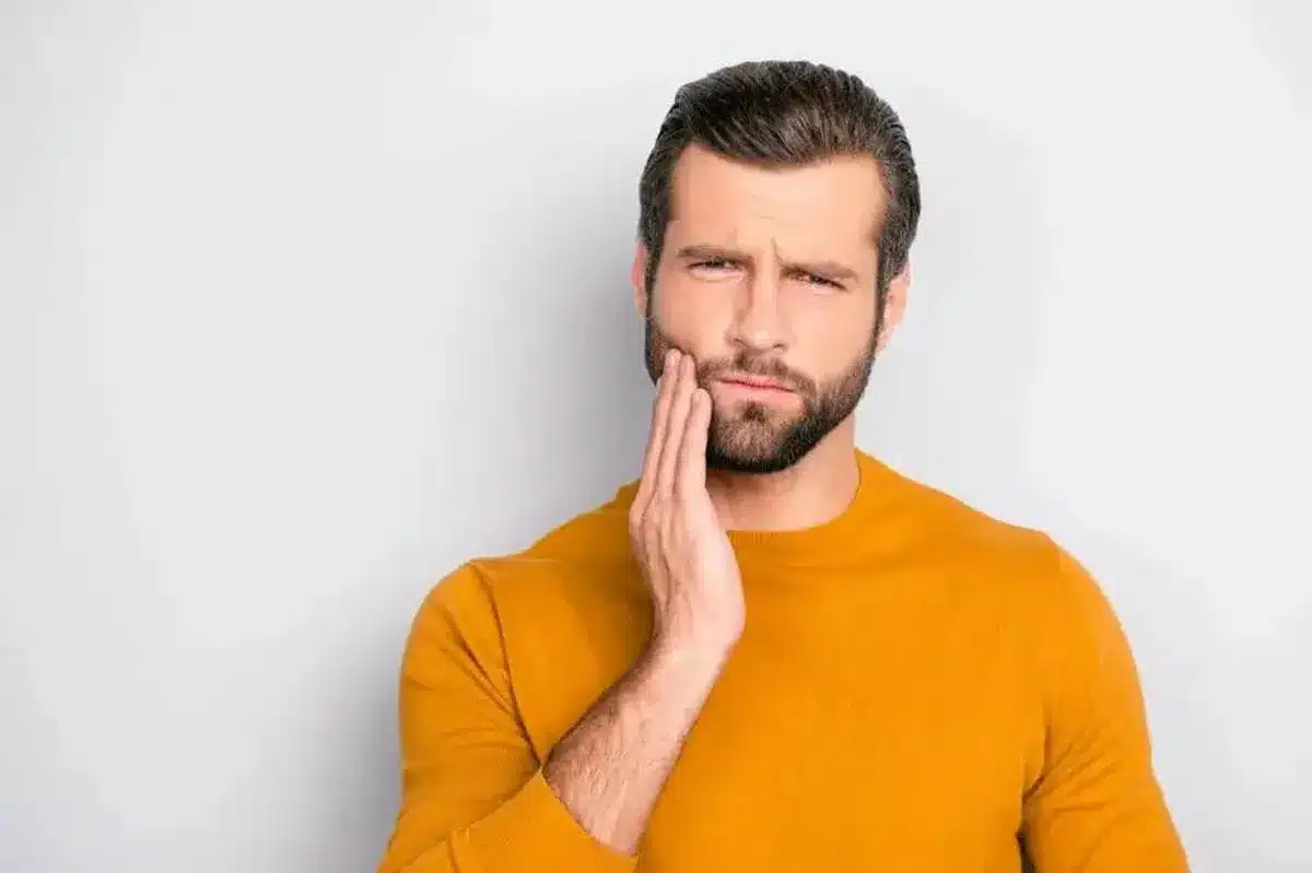Man in orange shirt touching cheek in pain against white background, dental discomfort concept.